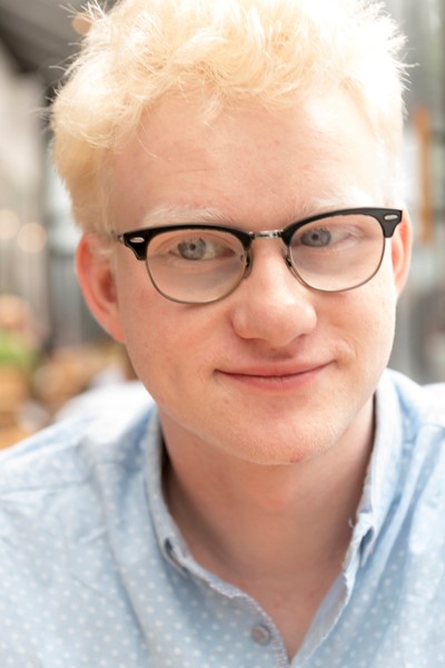 “Emmet Today” - a young man with short blond hair, light blue eyes, and glasses sits facing the camera with a confident yet understated smile on his lips