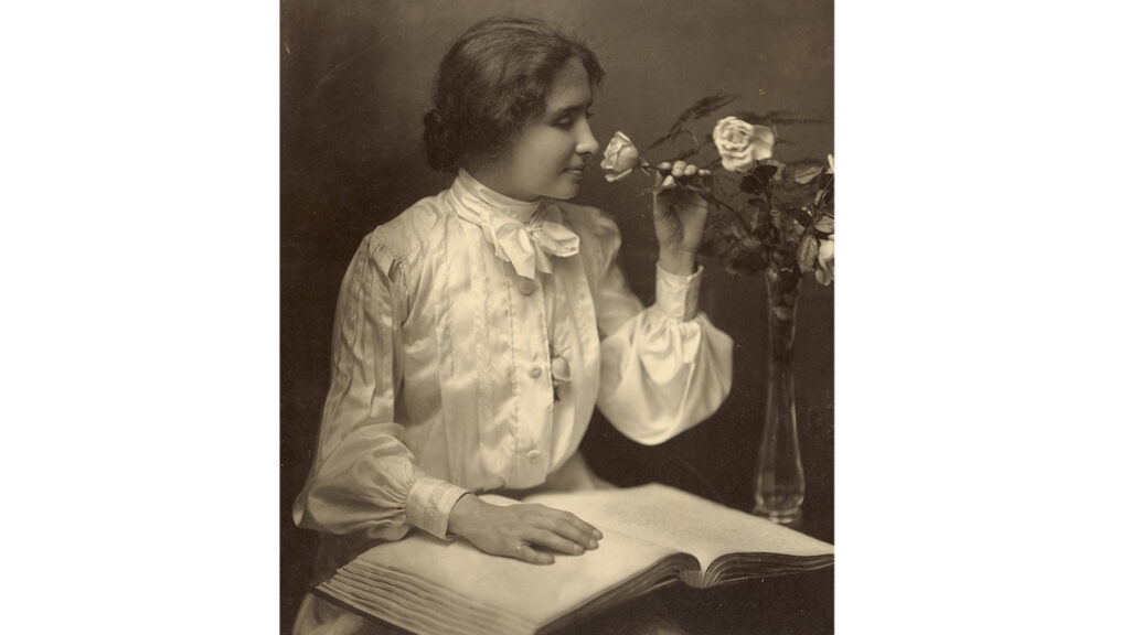 Helen Keller sitting at a desk with one hand on a braille book and the other hand holding a flower to her nose that she is smelling.  
