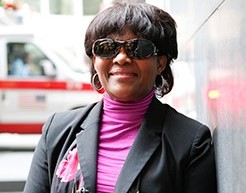 A woman standing outside with her white cane next to a building smiling at the camera