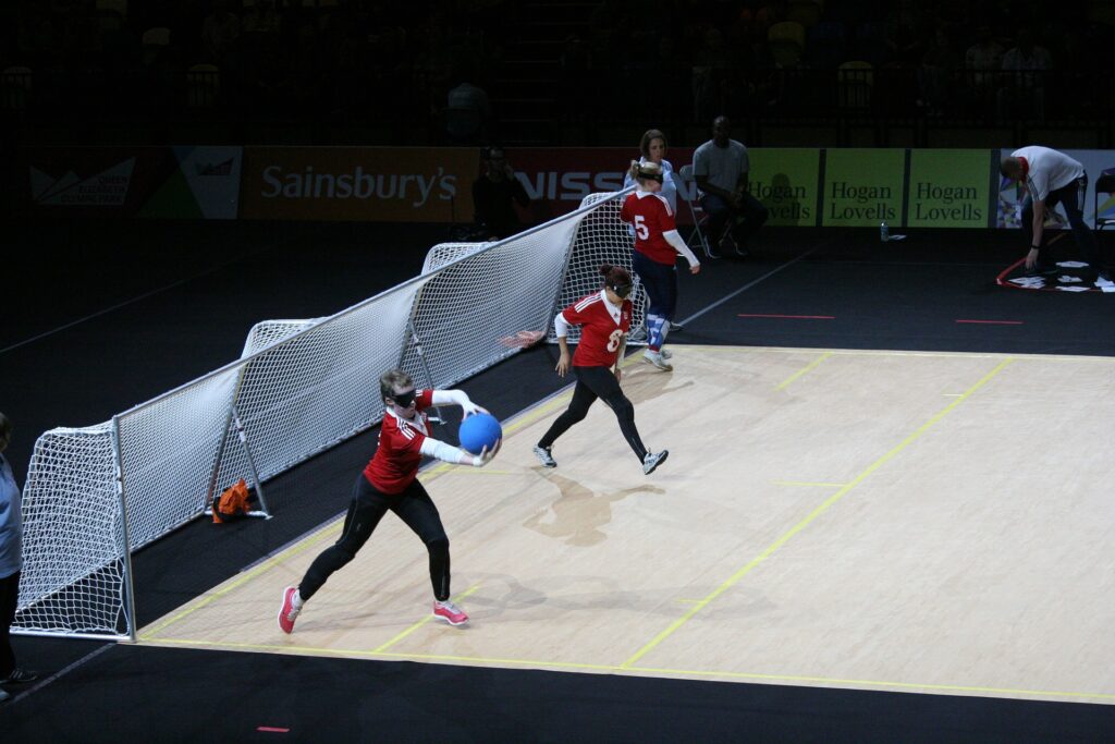 Three people with eyeshades and a uniform move on a goalball court, one prepares to roll a dodgeball-size ball.