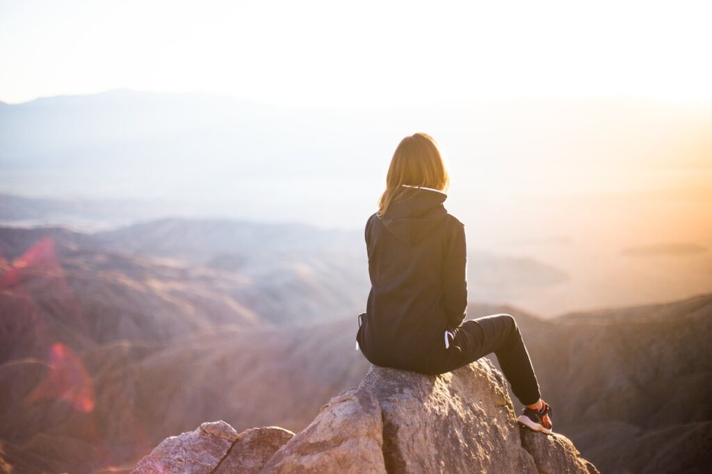 Young adult sits alone on a rock in a contemplative manner overlooking mountains 